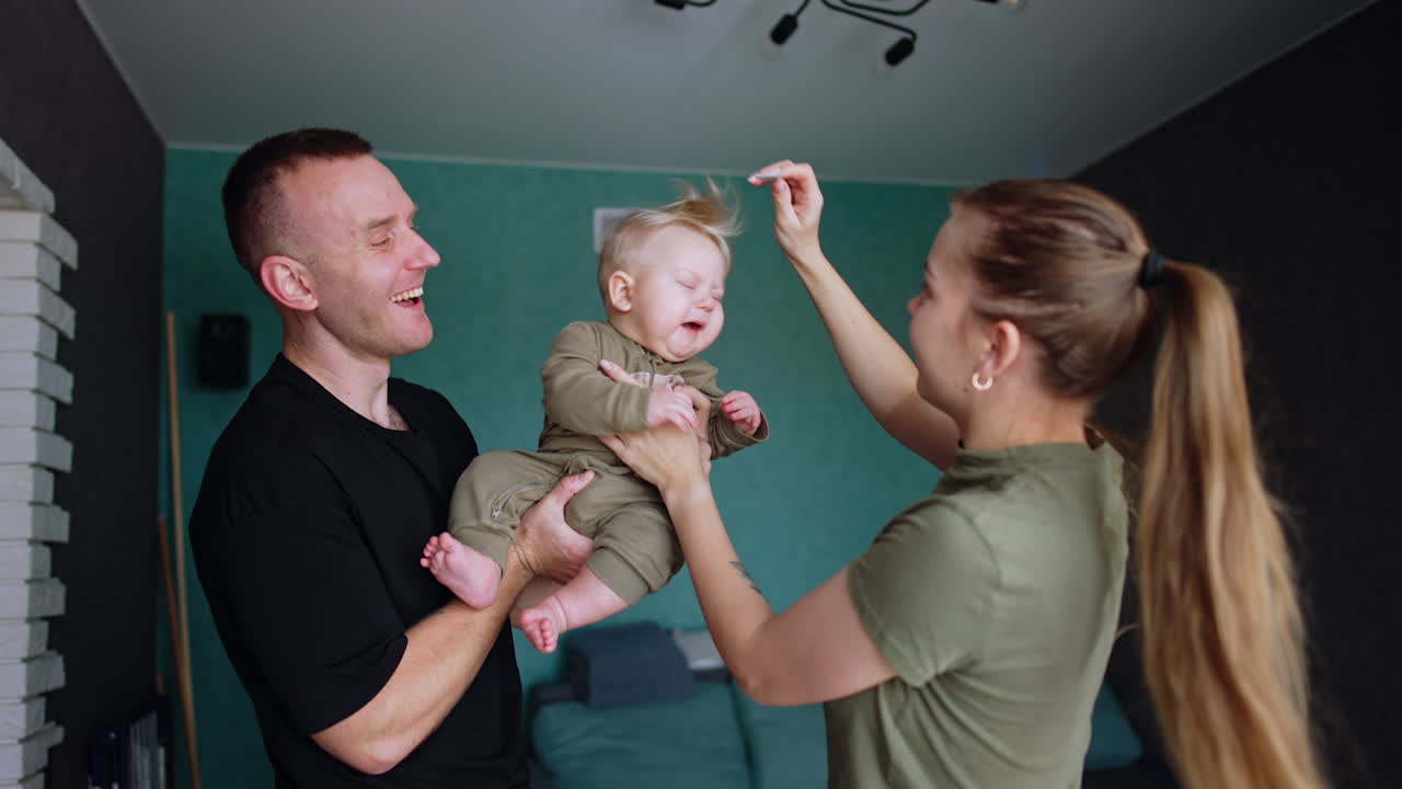 Man in black t-shirt holds a baby boy in hands. Mother tries to comb kid's hair and child doesn't like it.