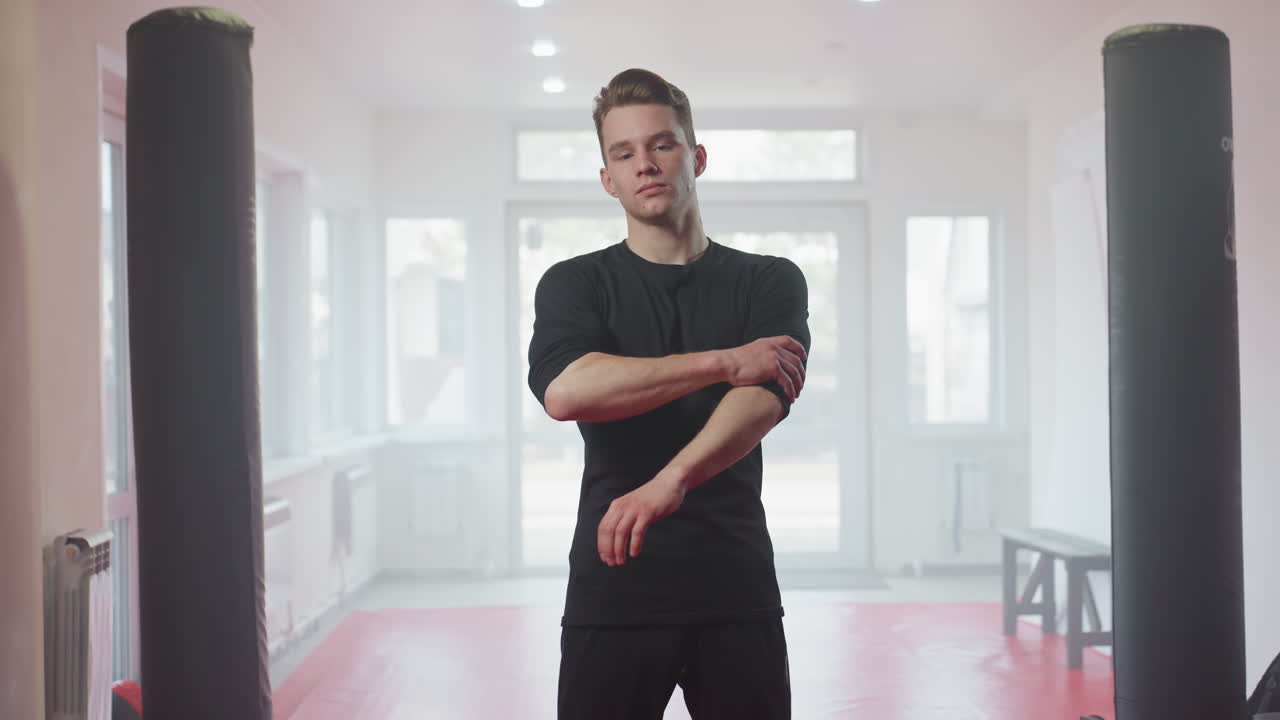 Athlete in black sportswear looks downward with serious thoughtful expression inside bright modern gym, standing near punching bag, representing focus, preparation before intense training