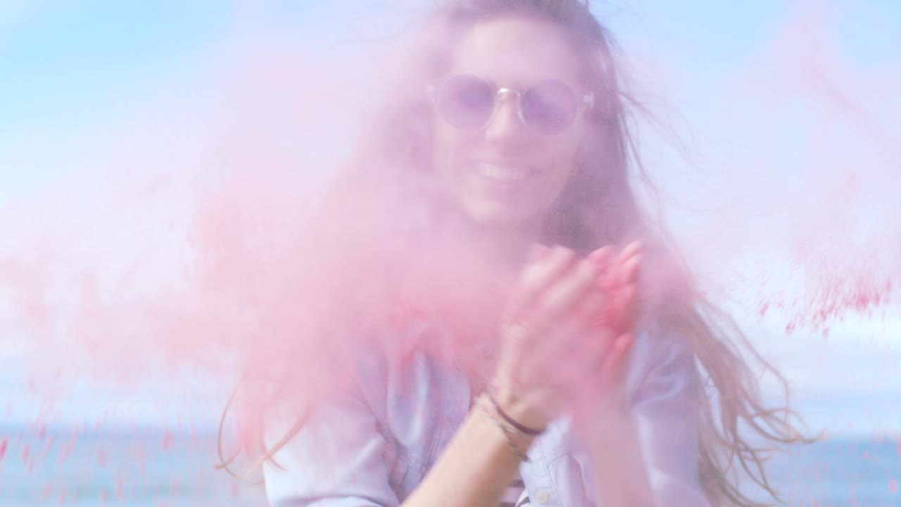 Bella giovane ragazza con capelli castani e occhiali da sole alla moda che soffia via la polvere colorata di Holi dalle mani e ride. Cielo azzurro limpido dietro di lei.