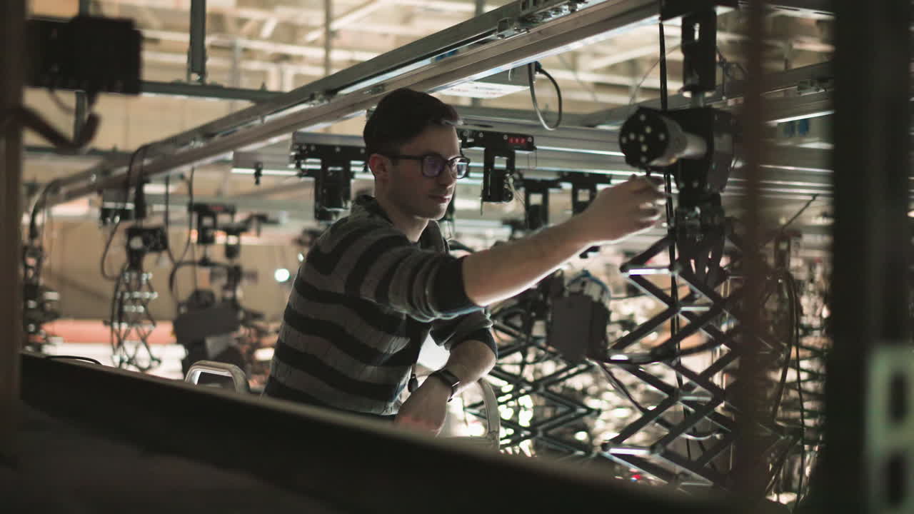 Technician adjusting studio lighting rigging equipment in overhead production space, focusing on rigging setup and fine-tuning lighting angle, crew working in background, professional studio environment