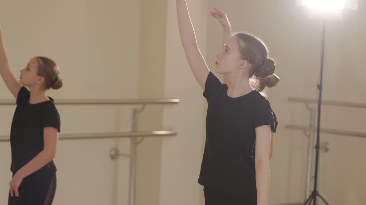 Un grupo de jóvenes estudiantes de ballet con ropa de baile negra practicando posiciones en un espacioso estudio de ballet con suelo de madera y barras de pared. Expresiones de concentración y movimientos sincronizados