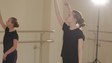 a group of young ballet students in black dancewear practicing positions in a spacious ballet studio with wooden flooring and wall-mounted barres. focused expressions and synchronized movements.