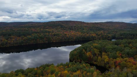 Aerial-captures-the-breathtaking-beauty-of-an-autumn-forest,-with-vibrant-orange-red-trees,-rain-filled-clouds,-and-the-tranquil-reservoir-reflecting-the-sky-like-a-mirror