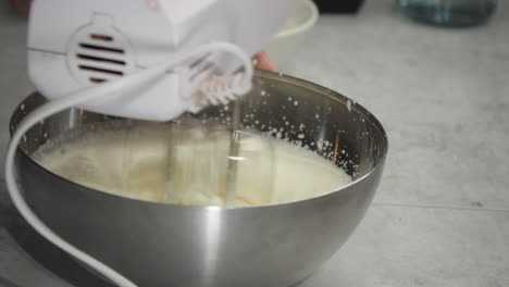 baker whisks cheesecake filling in metal bowl with hand mixer, closeup