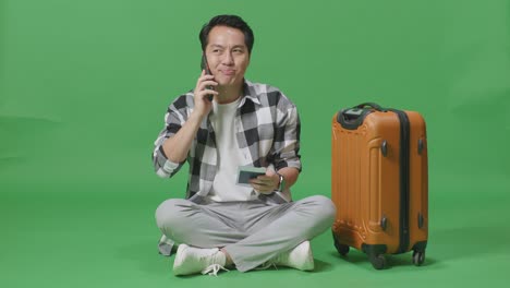 full body of asian male traveler with luggage and passport talking on smartphone while sitting in the green screen background studio