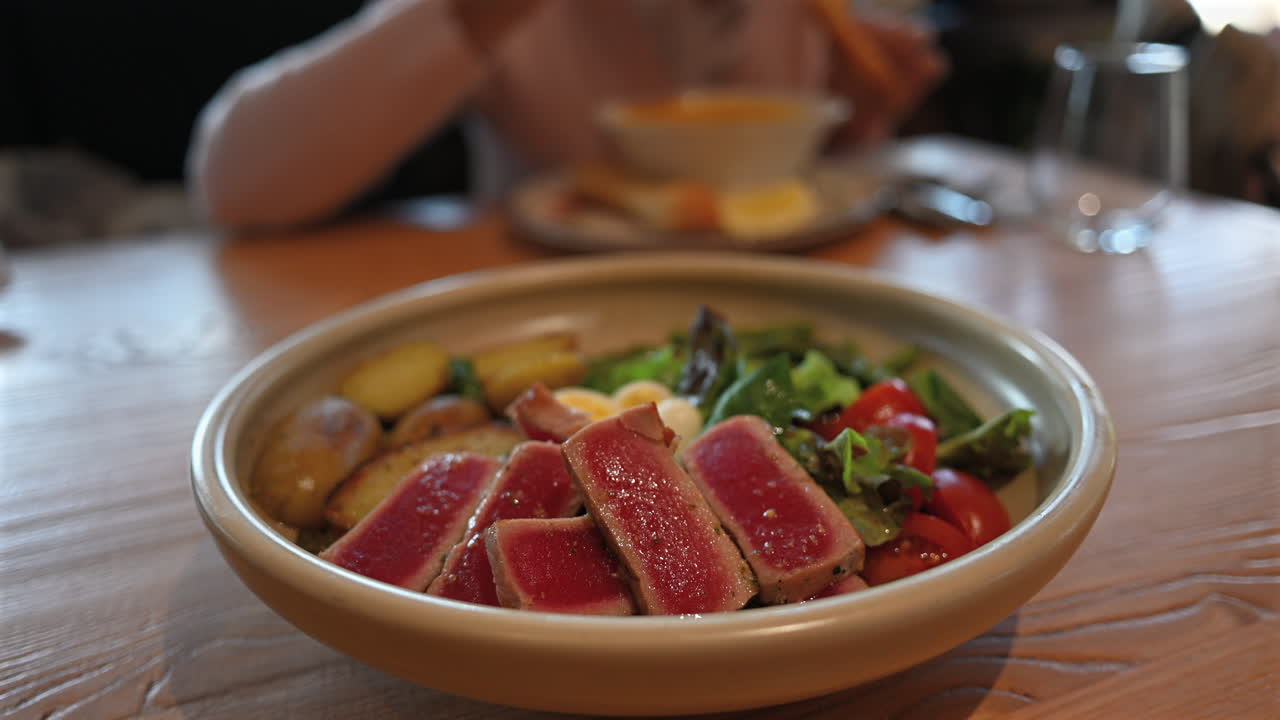 Fette di filetto di tonno scottato servite con patate, insalata fresca, pomodori e uovo su un tavolo di legno, con una donna che mangia sullo sfondo in un ristorante