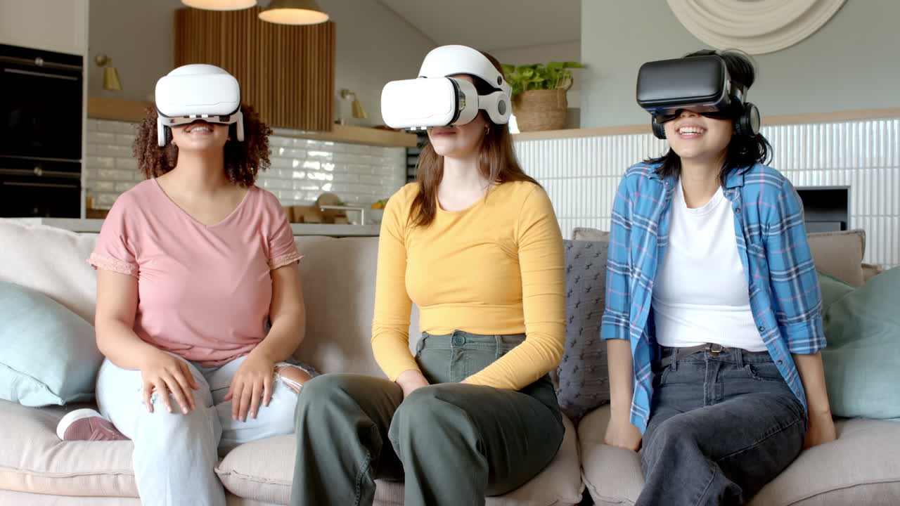 Wearing VR headsets, three diverse women friends sitting on couch enjoying virtual reality