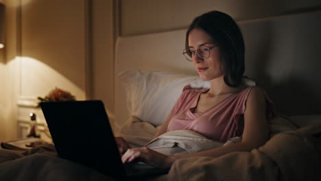 evening girl typing laptop working in bed. smiling relaxed woman browsing web