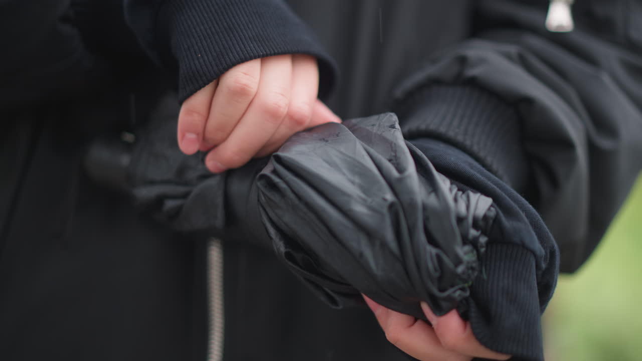 Packing wet umbrella into jacket hands, closeup of woman folding and securing compact umbrella, soft rain drains and droplets fall away, calm poststorm mood with neat movements and blurred green