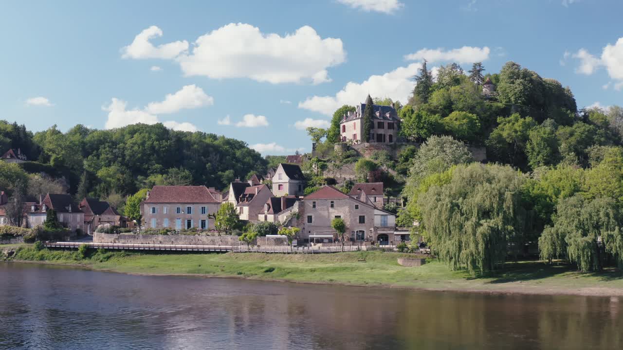 Splendida vista soleggiata del villaggio di Limeuil in Dordogna, Francia. Edifici storici in pietra si ergono su una rigogliosa collina verde, affacciati sul fiume tranquillo sotto un cielo blu con nuvole.