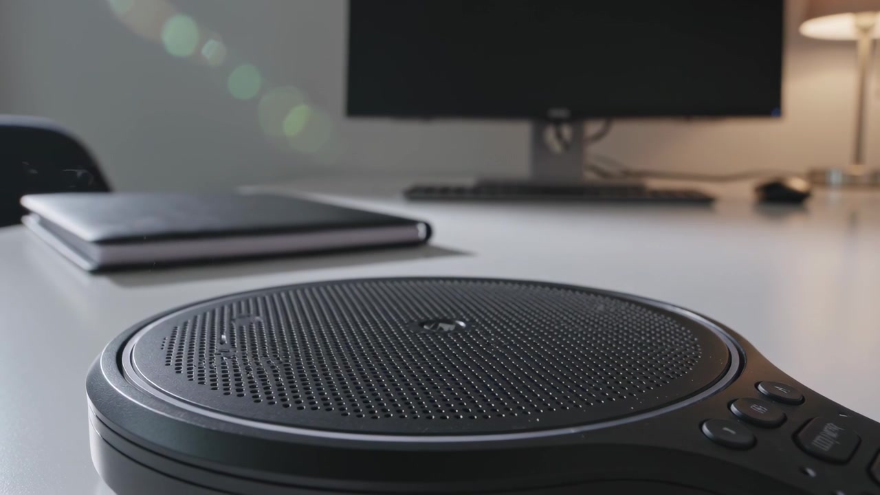 Recording camera capturing round speakerphone on white office desk, green lens-flare appearing left