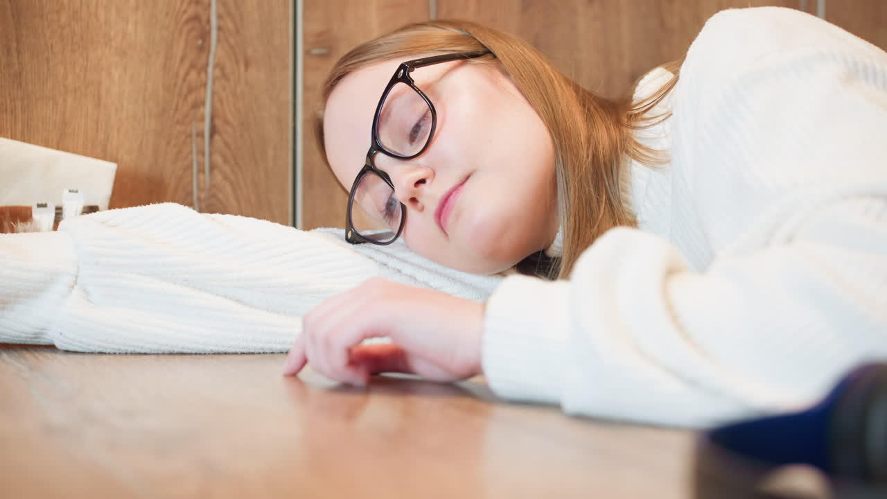 female student in white sweater lies with head on table, eyes closed and hand lightly tracing surface, appearing exhausted in quiet wooden setting with soft indoor lighting and napkin holder