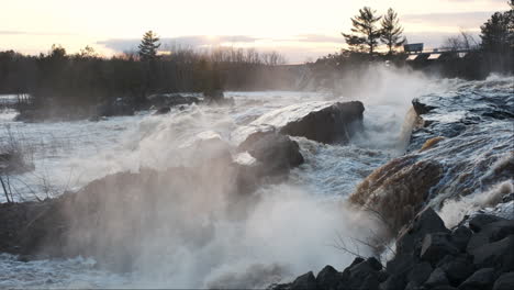 misty waterfalls cascade over rocks at sunset, the industrial silhouette of a hydropower plant in the background