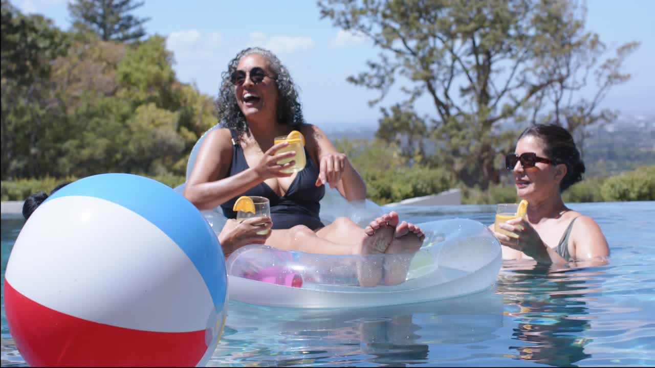 Beach ball drifting, leaning woman on float in swimsuit raising yellow cup, chatting in home pool