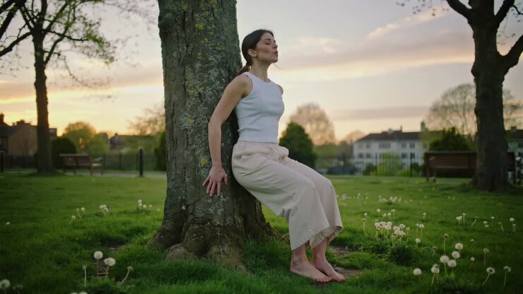Settling woman in white tank, wide pants practicing calm breathing in park, leaning on tree