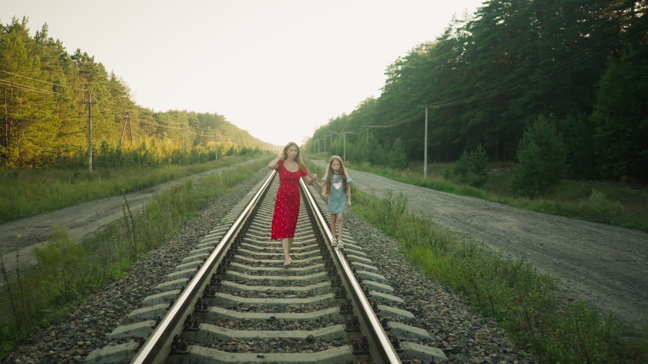 Lady in red dress walking barefoot on rail track adjusting dress while holding daughter hand as girl balances on beam during evening stroll in countryside surrounded by forest and gravel road