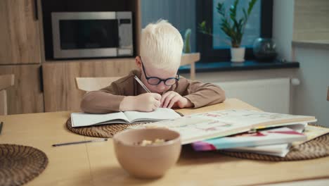 un niño albino con gafas azules escribe algo en su cuaderno y se prepara para el día siguiente en la escuela en casa en la cocina en un apartamento moderno por la noche