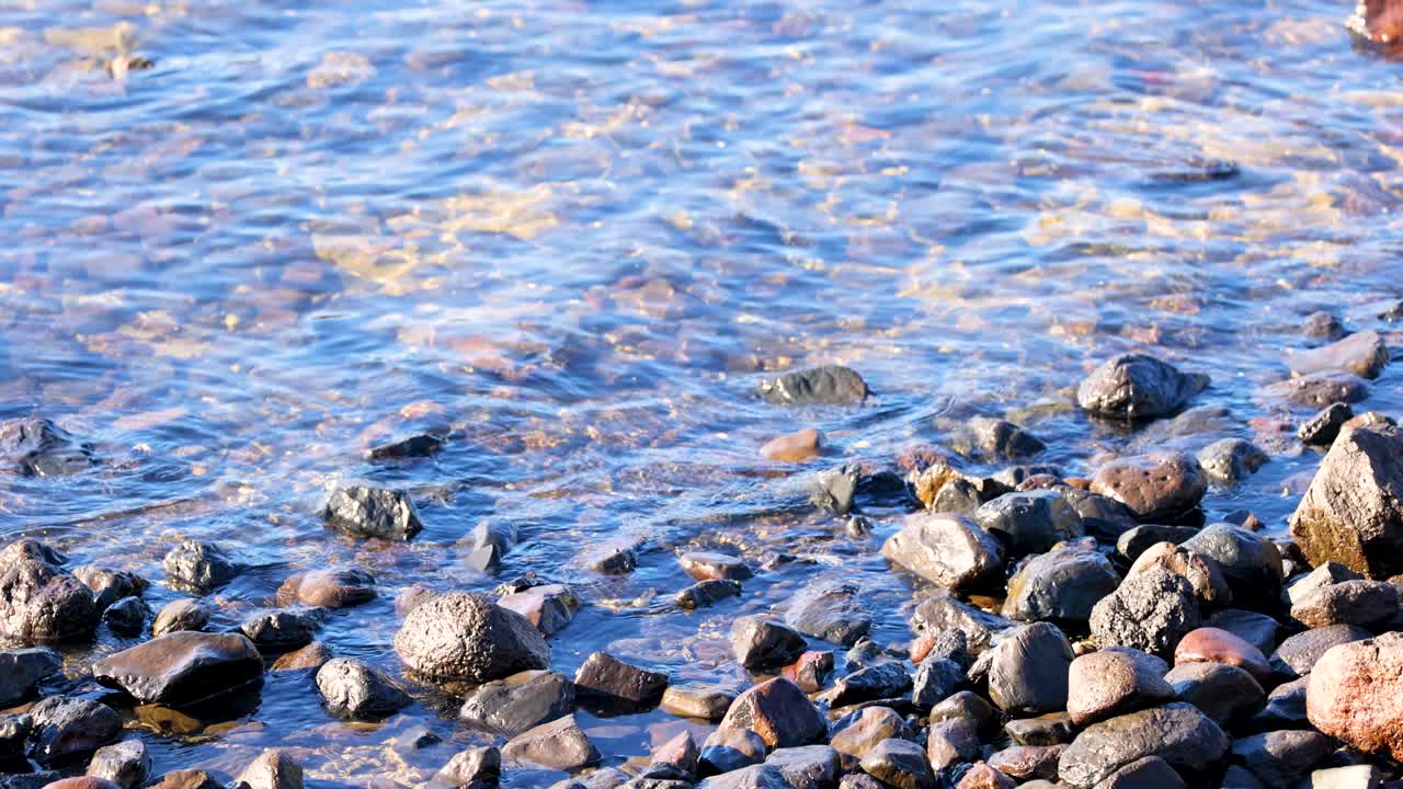 Waves gently lap against a rocky shoreline in Akaroa, New Zealand, under bright daylight, creating a serene coastal scene