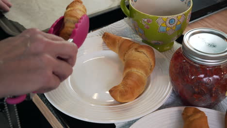 croissant being put on white breakfast plate with jam and cup by female hand using pink kitchen tongs