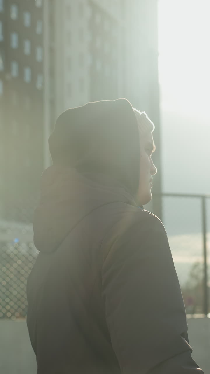 joven camina en la arena deportiva con rayos de luz brillando a su alrededor, con fondo con edificio residencial, coches estacionados y una valla