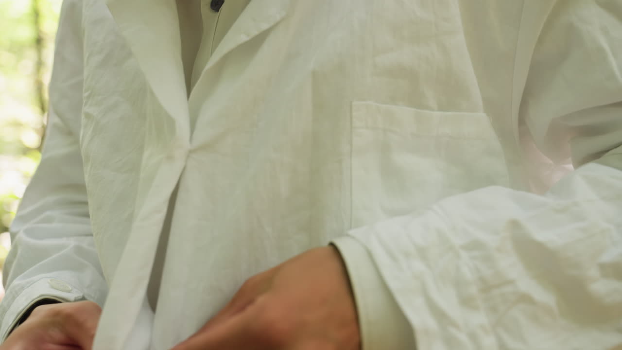 Close view of young botany student in white shirt buttoning coat in blurred forest with bright light, highlighting focus, outdoor preparation, and lifestyle detail surrounded by natural greenery
