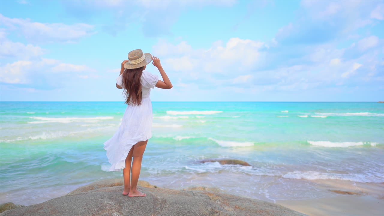 한 젊은 태국 여성이 열린 바다 앞 해변의 바위 가장자리에 서서 팔을 위로 올리고, 슬로모션 주간 태국