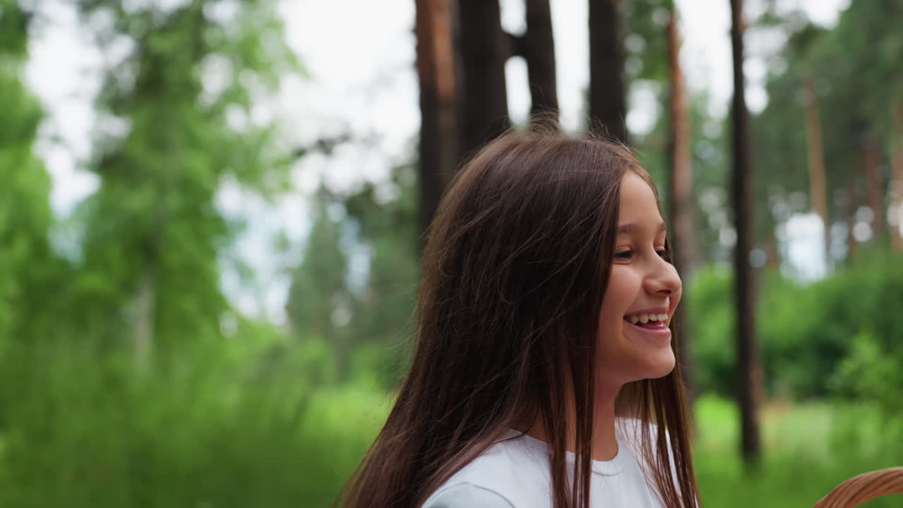 vista media de una chica alegre que pasea por un sendero del bosque con una cesta de picnic, levantando suavemente la toalla del interior con entusiasmo, disfrutando del aire fresco, la vegetación, la luz del sol y el ambiente tranquilo