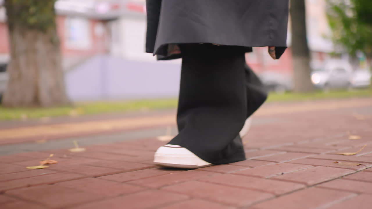Close-up of woman in black coat walking along city sidewalk in white sneakers, autumn leaves scattered on pavement, urban lifestyle moment with movement and atmosphere in calm seasonal outdoor environment