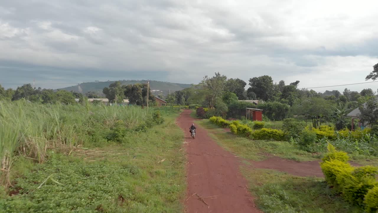 seguimiento de tomas aéreas frente a un hombre africano con casco en una motocicleta que recorre un camino de tierra a través de un pueblo rural