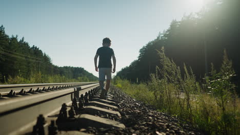 back view of young boy wearing dark shirt and light shorts walking carefully on rail track ballast, balancing step by step as sun glow filters through tall trees in quiet forested rural setting