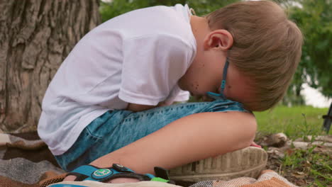 a child is seated on a mat with head bowed down, resting against his knees near a tree trunk, there is a partial view of a bicycle in the background