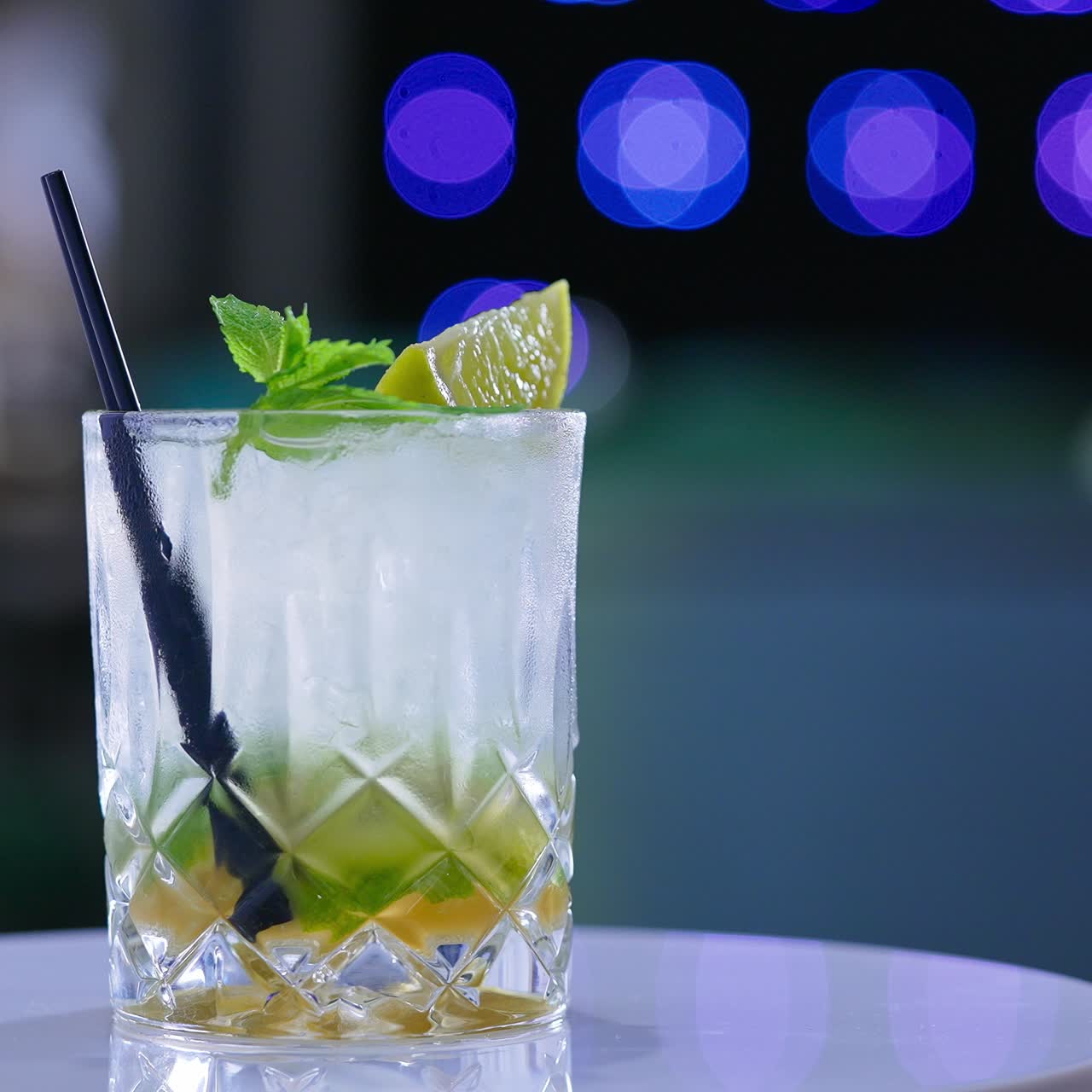 Misted glass full of beverage with two short black straws in it. Rotating glass with lemon and mint leaves on top. Close up. Blurred backdrop