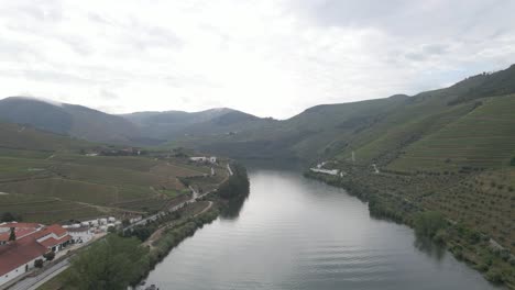 Vista-Aérea-De-La-Ciudad-Vinícola-De-Pinhão-Portugal,-Drone-Avanzando-Sobre-El-Río-Duero-Mostrando-Las-Plantaciones-De-Vides-Y-Las-Montañas-Al-Fondo