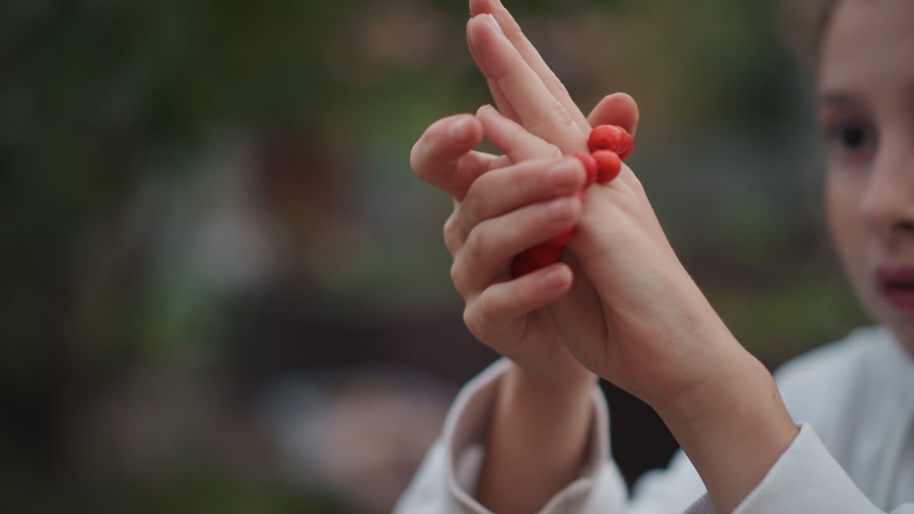 Hands threading red beads on finger, young girl practicing dexterity outdoors closeup, sliding round beads along string and finger playful curiosity, focused expression, shallow depth of field, warm
