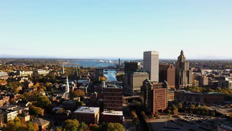 aerial fly over above the high rise buildings following the moshassuck and providence rivers in downtown providence, rhode island with smokestacks, bridges, wind turbines and church steeples beyond.