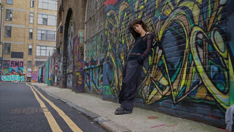 full length outdoor fashion portrait of young alternative style woman leaning against graffiti covered wall in london city street uk in real time