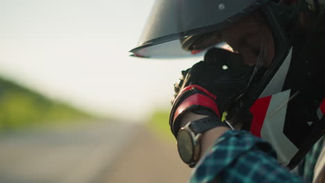 primer plano de la mano de un motociclista cubriendo su cara a través de la visera del casco en una tranquila carretera rural, ella usando un guante de protección y un reloj inteligente