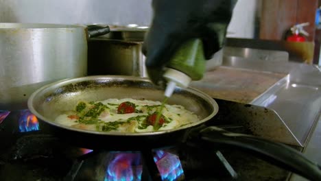 chef wearing black rubber gloves pours green mixture into cooking pan with white sauce and tomatoes