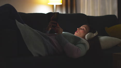 Woman Wearing Wireless Headphones Lying On Sofa At Home At Night Streaming Or Looking At Online Content On Mobile Phone 4