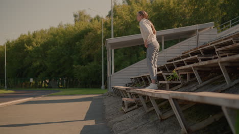 lady casually walking up rustic stadium seating in relaxed, natural posture, dressed in jeans and hoodie, background includes empty stadium bleachers, electric poles, and lush green trees