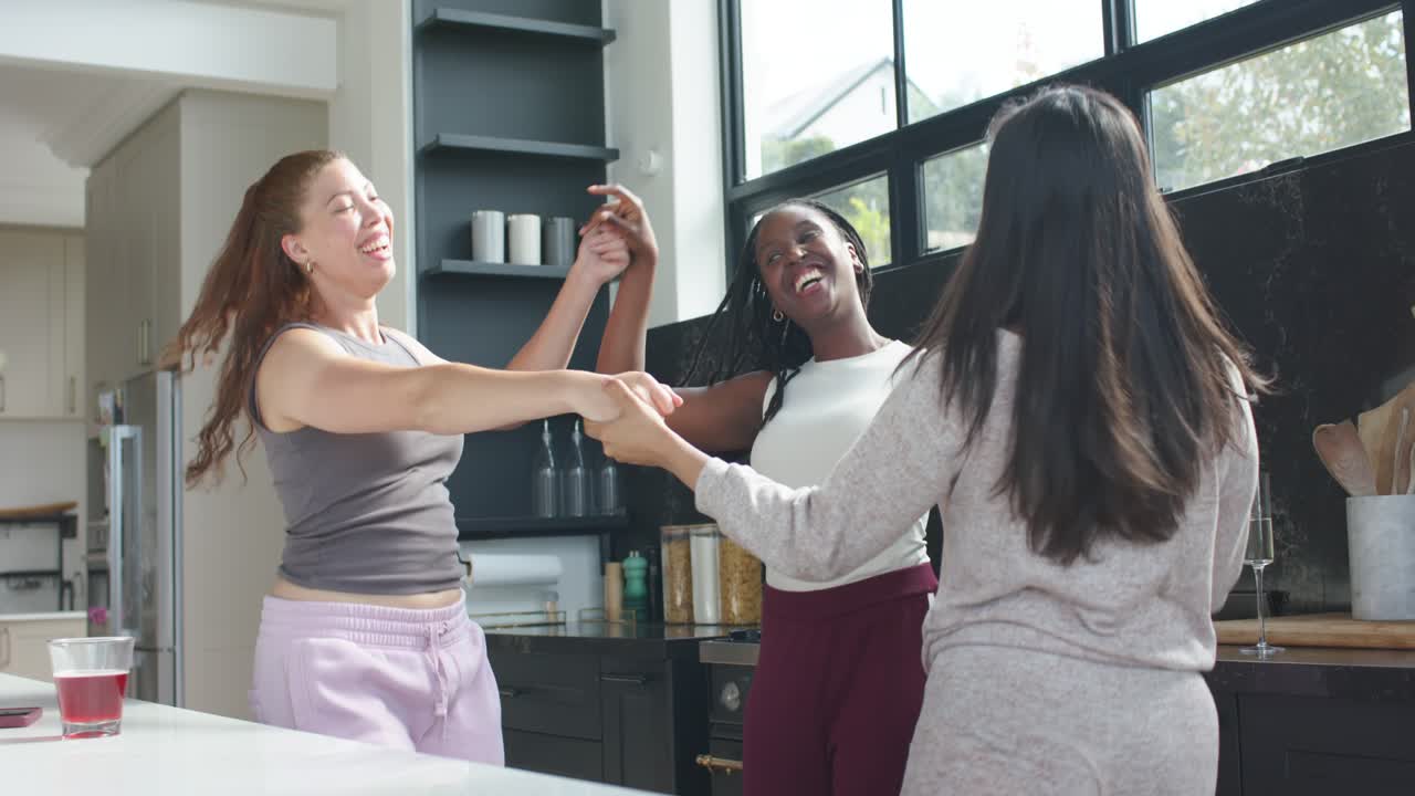 Diverse female friends linking hands, spinning in kitchen near counter with red drink celebrating