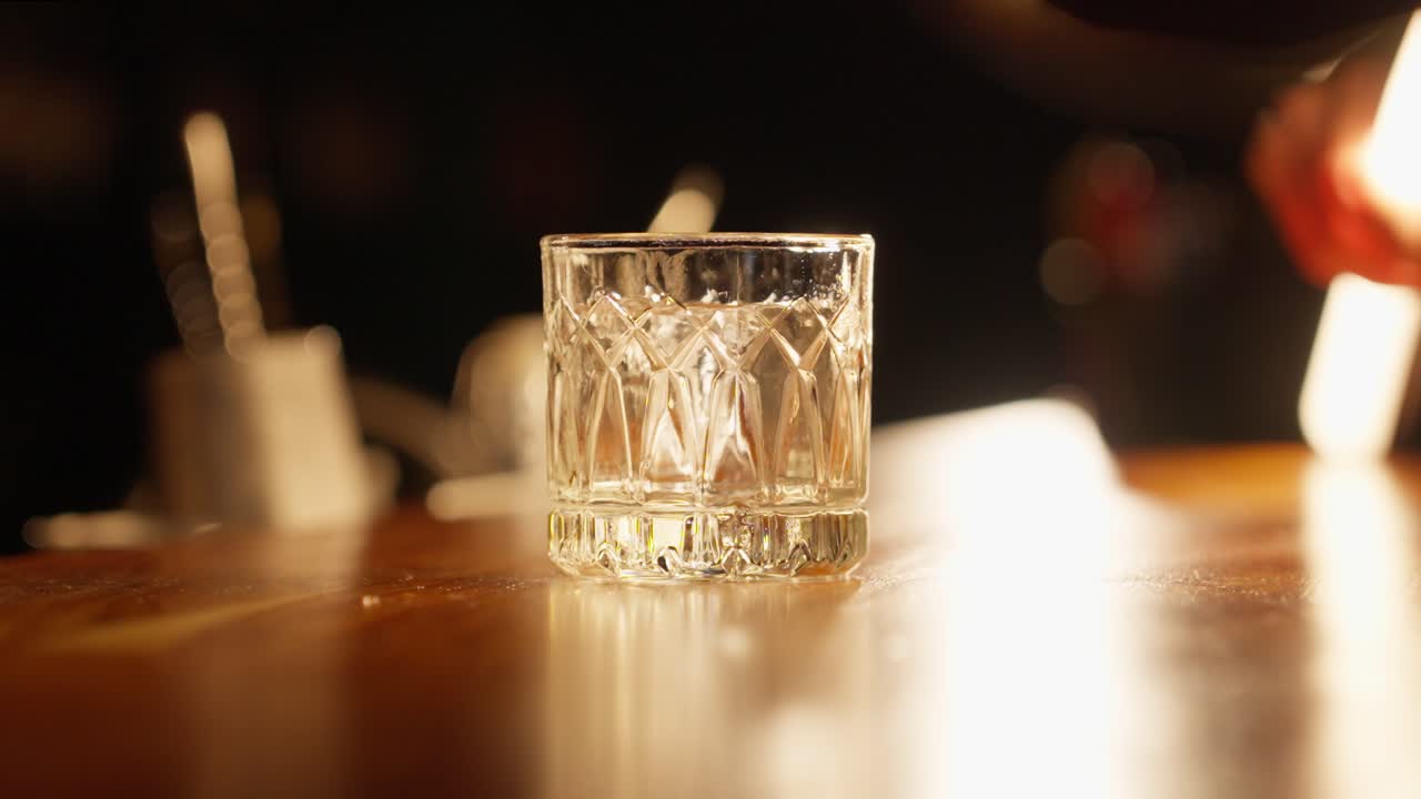 A close-up of a professional bartender's hands using tongs to spin a hand-carved, clear ice cube in a crystal rocks glass to chill the vessel at a bar