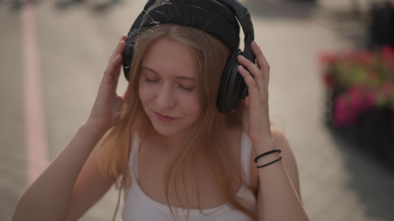 ragazza caucasica in una piazza cittadina che si sistema le cuffie, annuisce a ritmo di musica e sorride, accennando passi di danza e chiudendo gli occhi, stile con berretto, luce del sole pomeridiana con bagliore tenue, artista spontanea