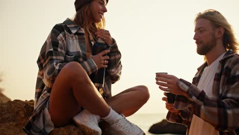 feliz chica rubia en una camisa a cuadros vierte té caliente de un termo negro a su novio rubio con barba en una taza especial, sentada en una piedra grande en una orilla rocosa cerca del mar por la mañana