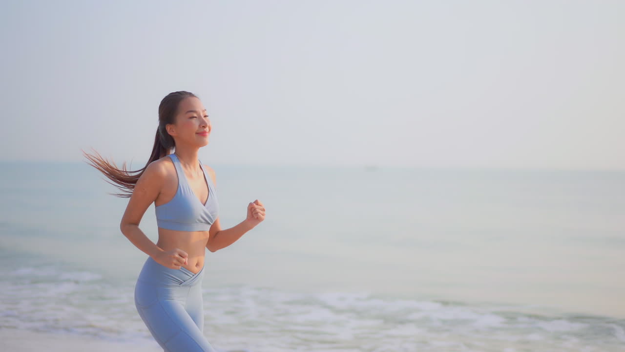 Slow-motion of a fit, young, Asian woman dressed in activewear runs up the beach while the waves roll in on the beach. copy space