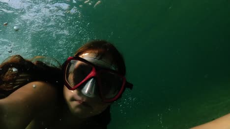 underwater selfie of young red-haired girl with diving mask holding camera while swimming in transparent sea water