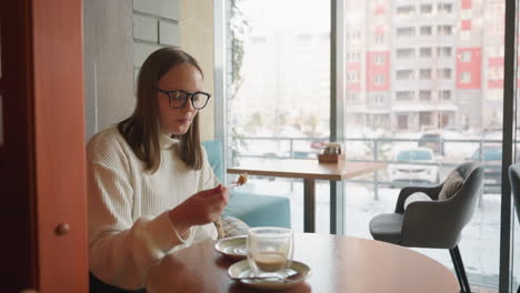 girl in glasses enjoying dessert with fork while seated at round table near large window inside cafe, outdoor urban scenery with parked cars and snowy buildings visible through glass