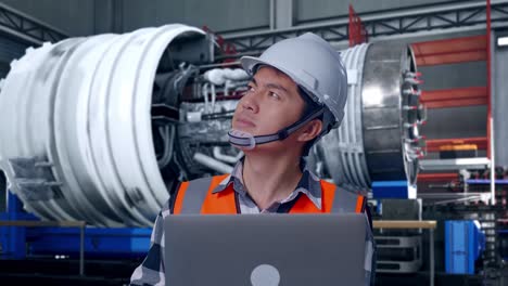 close up of asian male engineer with safety helmet working on a laptop and looking around while standing with airplane engine maintenance conducted, analytics checking the turbine