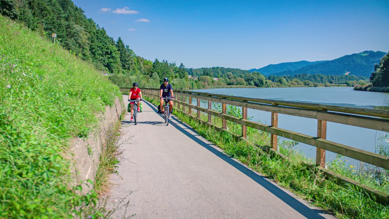 Casual couple enjoys a bike ride on a path on the edge of a lake in lush green landscape. Slow Motion shot