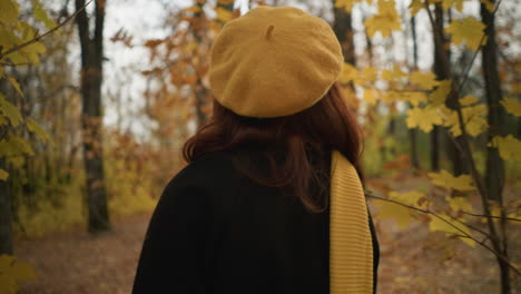 back view of lady in yellow beret and muffler strolling through forest, gently shifting dry branches aside to pass, gazing left as golden autumn leaves sway in soft breeze